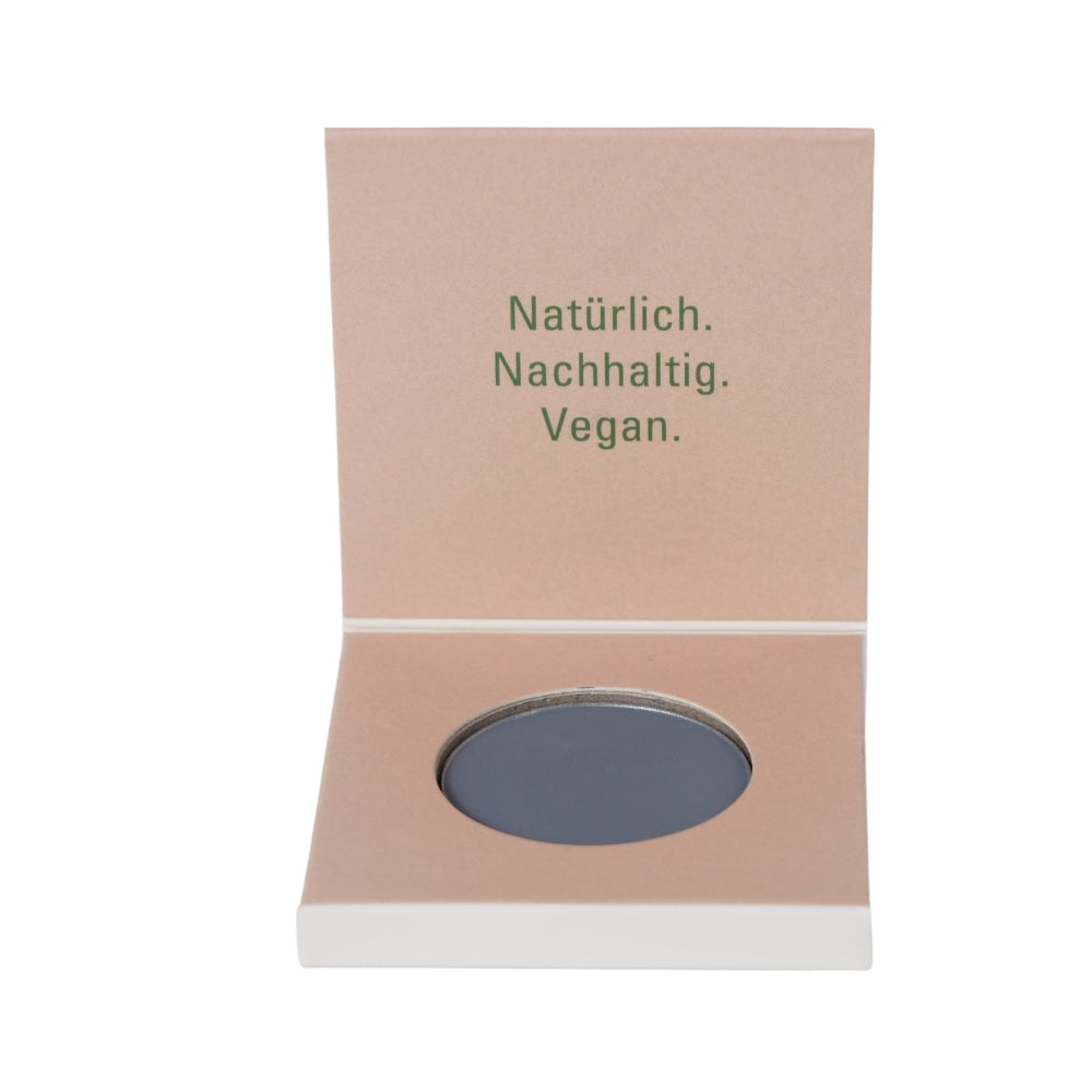 Open beige cardboard packaging reveals a round black cosmetic labeled Eyeshadow Blue Mystery MATT. The inside lid reads: Natürlich. Nachhaltig. Vegan. The packaging exudes a natural and sustainable theme, perfect for effortlessly applying mineral eyeshadow with grace.