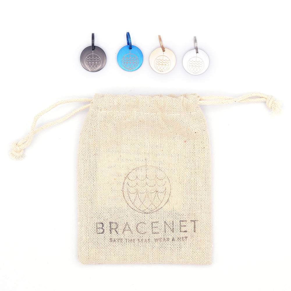 Vier Schlüsselanhänger 1 Anhänger Maritim von Bracenet ruhen über einem beigen Kordelzugbeutel mit Bracenet-Logo. Diese stilvollen Schlüsselanhänger aus Edelstahl unterstützen ebenfalls die Mission „Meere schützen“.