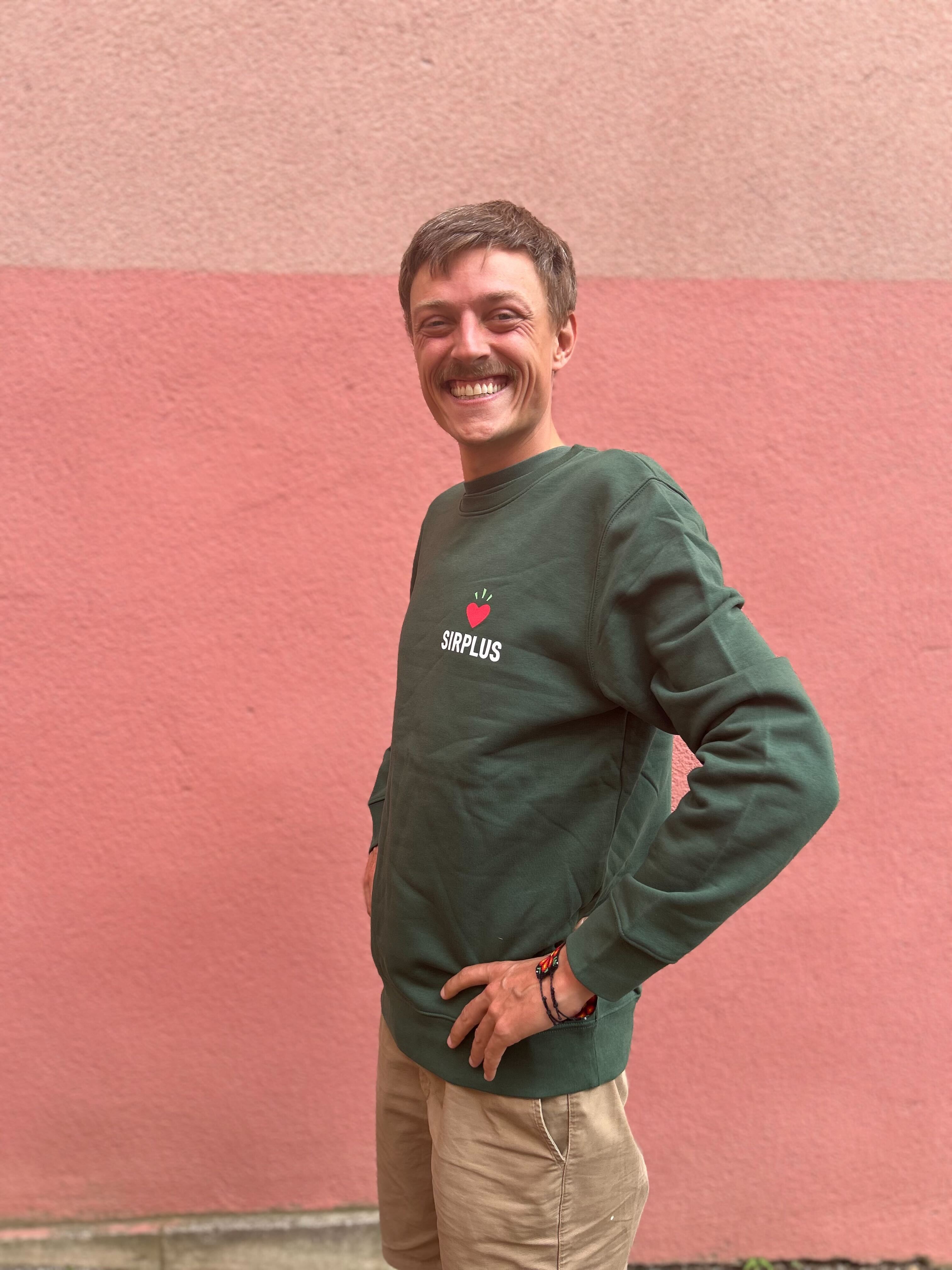 Eine lächelnde Person in einem grünen SIRPLUS-PULLOVER und beigen Hosen steht vor einer zweifarbigen rosa Wand.