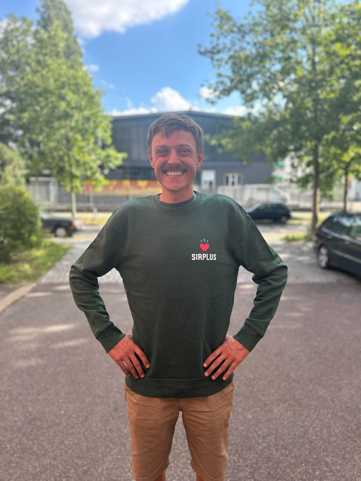 Eine lächelnde Person steht draußen mit den Händen in den Hüften, in einem grünen SIRPLUS PULLOVER aus Bio-Baumwolle, vor einer ruhigen Kulisse aus Bäumen und Himmel.