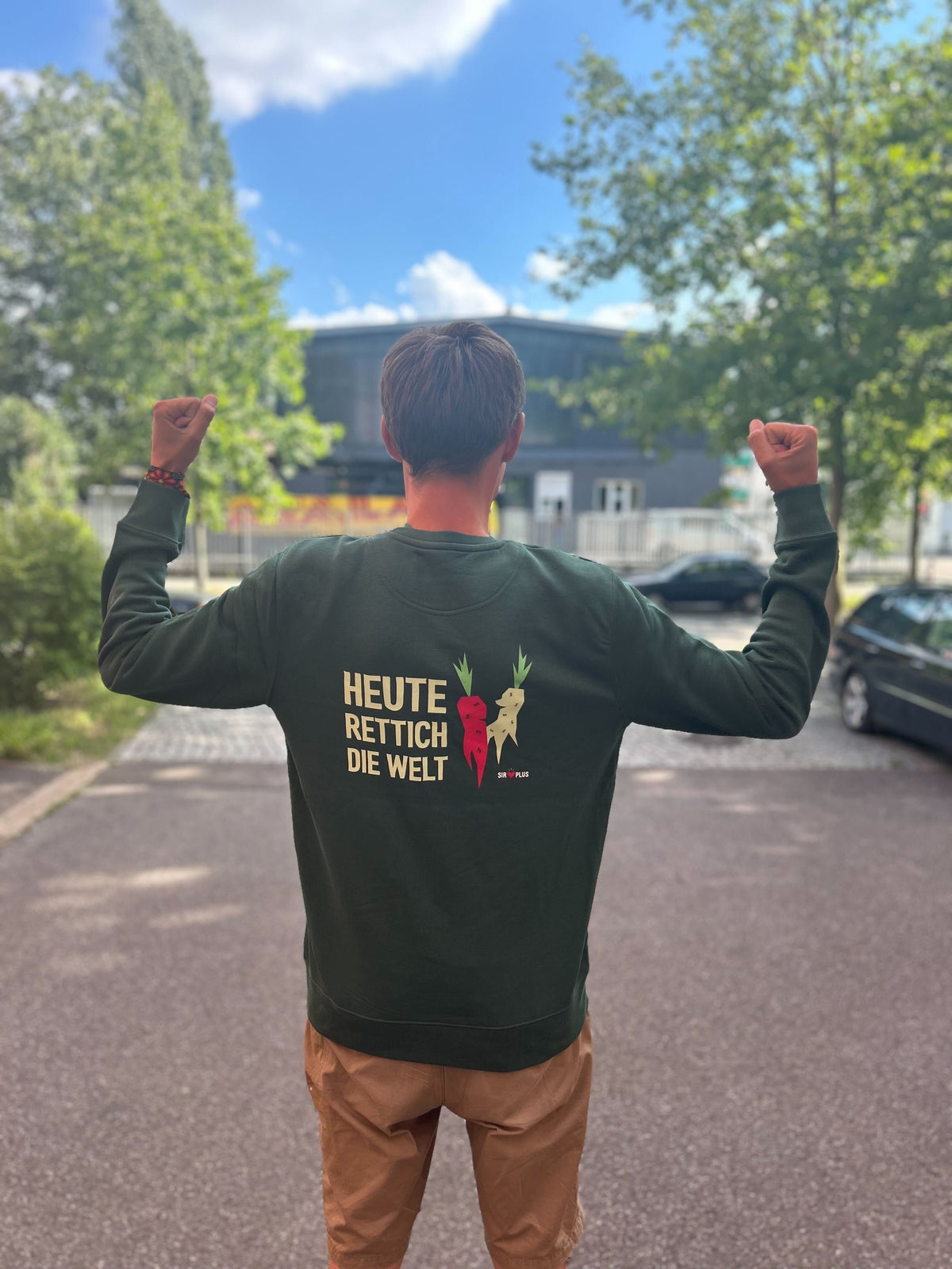 Eine Person mit erhobenen Armen steht auf einer sonnigen Straße und trägt den SIRPLUS PULLOVER aus Bio-Baumwolle. Er trägt eine inspirierende Botschaft und eine Karottengrafik.