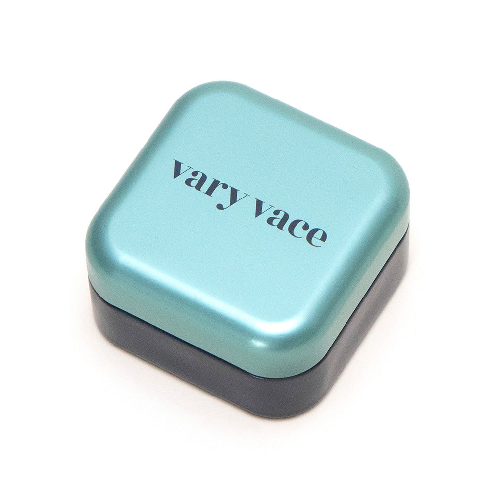 Der Lidschatten Diane von Vary Vace wird in einer blaugrünen, quadratischen Blechdose mit der Aufschrift „Vary Vace“ auf dem Deckel geliefert, die einem perlweißen Farbton ähnelt, der mit silbernen Glitzerpartikeln verziert ist.