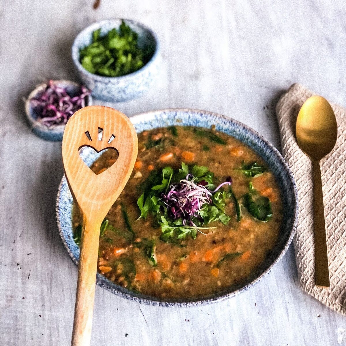 Eine Suppenschüssel mit dazugehörigem SIRPLUS Kochlöffel, gefertigt aus hochwertigem Buchenholz, steht mit kleinen Kräuterschälchen und einem goldenen Löffel auf einer Serviette.