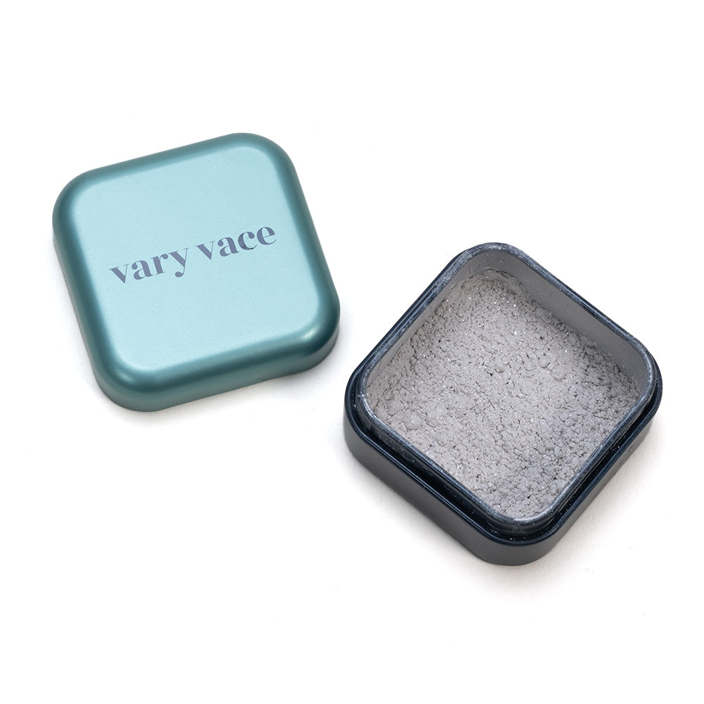 Eine elegante offene quadratische Dose von Vary Vace gibt den Blick auf Eye Shadow Diane frei, ein graues Puder, das mit silbernen Glitzerpartikeln versetzt ist, neben einem Deckel, der an holografischen Lidschatten erinnert.